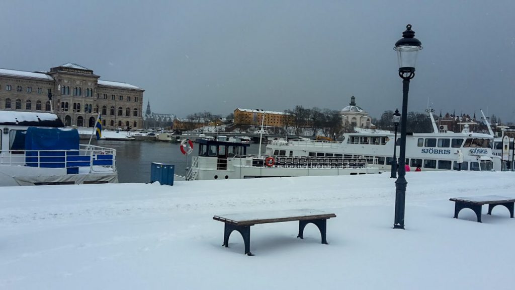 Bänk, snö, båtar och Skeppsholen i Stockholm.